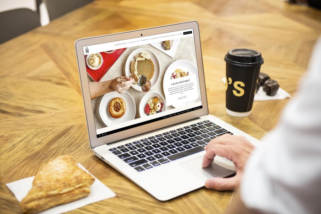 A person is using a laptop on a wooden table, which displays a restaurant website featuring various food dishes. A pastry on a napkin and a black coffee cup sit next to the computer.