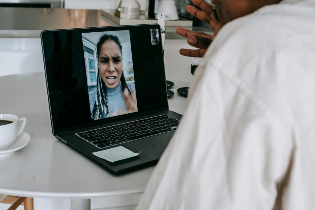 A person in a light-colored shirt is seen from behind, gesturing during a video call on a laptop.