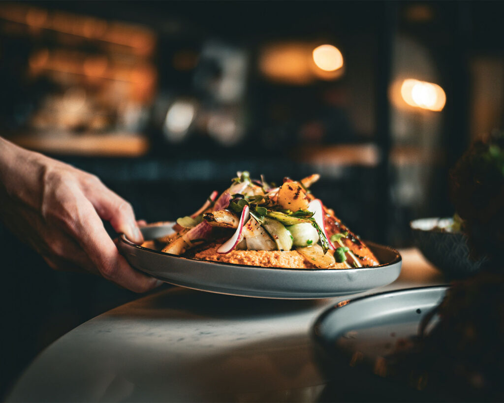 A hand gently places a freshly prepared dish onto a wooden table, suggesting food service or meal presentation in a dining setting.