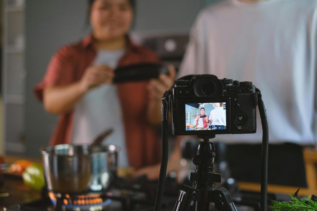 A professional camera on a tripod captures two people preparing a meal in a kitchen. The foreground focuses on the camera's digital display.