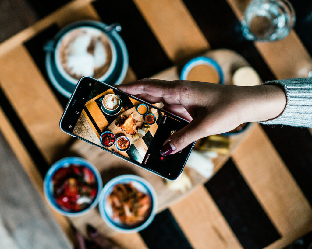 Hand holding a smartphone above a dining table, capturing a photo of a beautifully plated meal in natural café lighting.