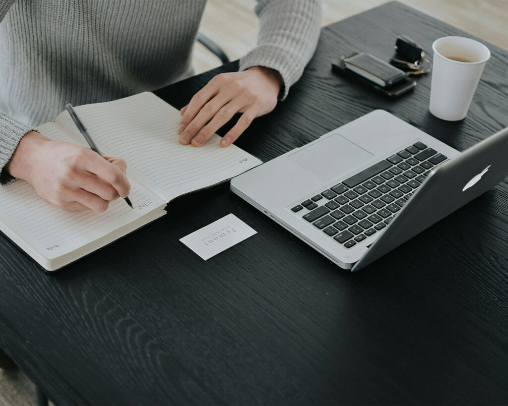 Close-up of a person’s hand writing notes in a notebook, with an open laptop beside her, suggesting focused study or work
