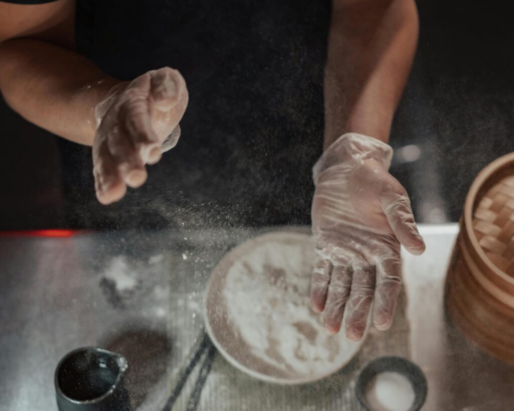 A person wearing clear plastic gloves claps their hands together, sending a fine cloud of white flour into the air.