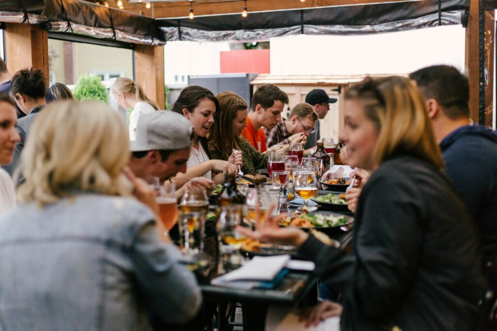 A large group of friends is gathered at a long outdoor table, enjoying a communal meal and drinks under a canopy with string lights.