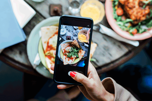 A person holds a smartphone over a wooden table to take a photo of a plated meal featuring wraps and greens. The table is filled with various dishes and drinks, captured