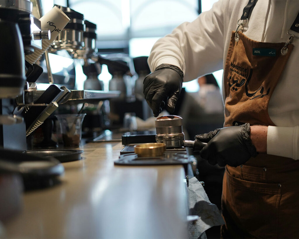 A barista carefully brewing coffee using an espresso machine, with steam rising and warm lighting highlighting the café’s craft and attention to detail.