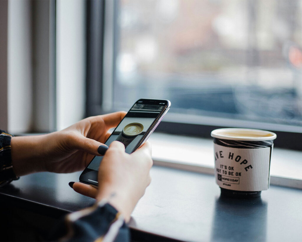 A hand holding a phone while photographing a cup of coffee on a table, capturing a casual café moment