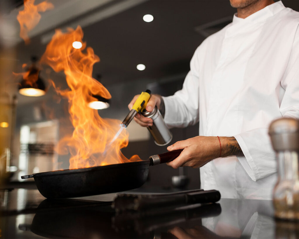 A professional chef in a busy kitchen flambéing a dish, flames rising dramatically from the pan. The chef’s focused expression and skilled hand movements highlight the artistry of cooking.