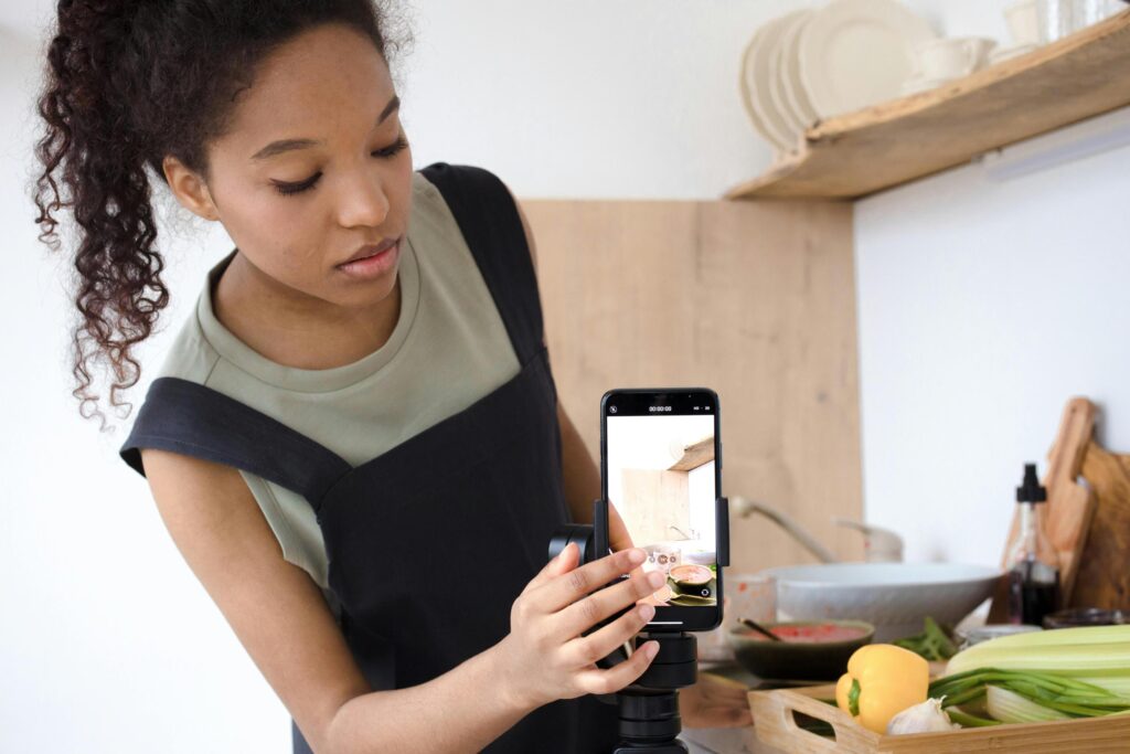 A woman with curly hair stands in a kitchen, carefully adjusting a smartphone mounted on a tripod to film a cooking demonstration.