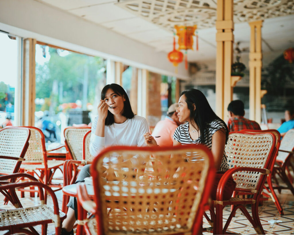 A group of diners sitting at a restaurant table, laughing and smiling as they taste their meals. The scene conveys warmth, enjoyment, and the social aspect of sharing food.