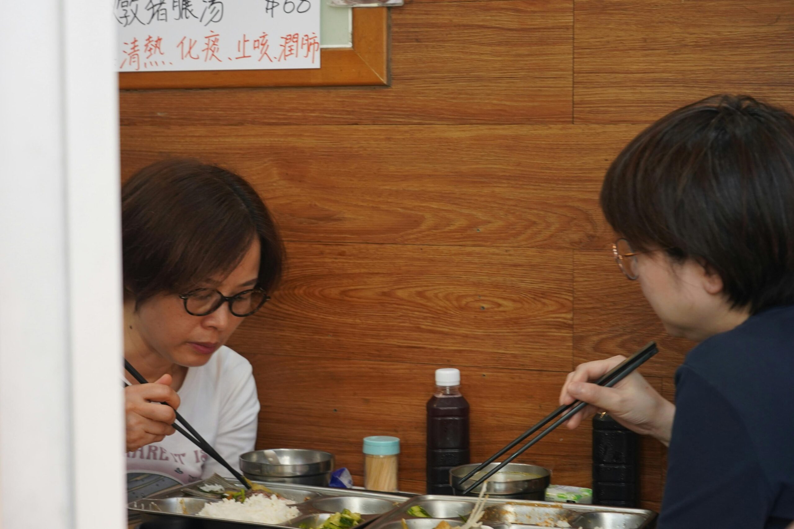 Two people wearing glasses sit at a wooden table, using chopsticks to eat from divided metal cafeteria trays. A handwritten sign in Chinese characters is posted on the wood-paneled wall.
