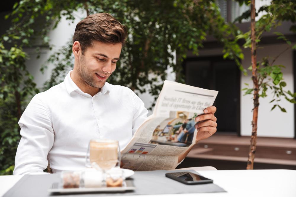 A smiling man in a white shirt sits at an outdoor table while reading a newspaper. He has a coffee and a smartphone nearby, with lush green trees and a building in the background.