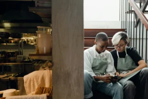 Two workers wearing matching light green aprons sit together on a wooden staircase while looking intently at a smartphone. The scene is captured from a side angle, revealing a glimpse of a commercial kitchen.