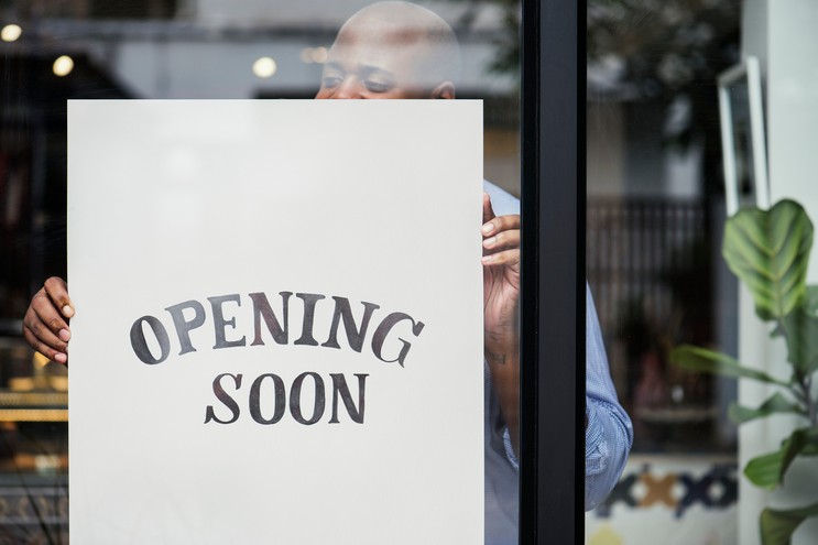 A man is shown through a glass window as he hangs a large white sign that reads "OPENING SOON" in bold black letters. The background reveals a glimpse of a modern, bright interior, suggesting a new storefront or restaurant preparing for its debut.