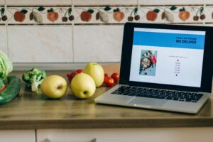 A laptop displaying a food delivery website sits on a kitchen counter next to fresh produce like apples, broccoli, and tomatoes.