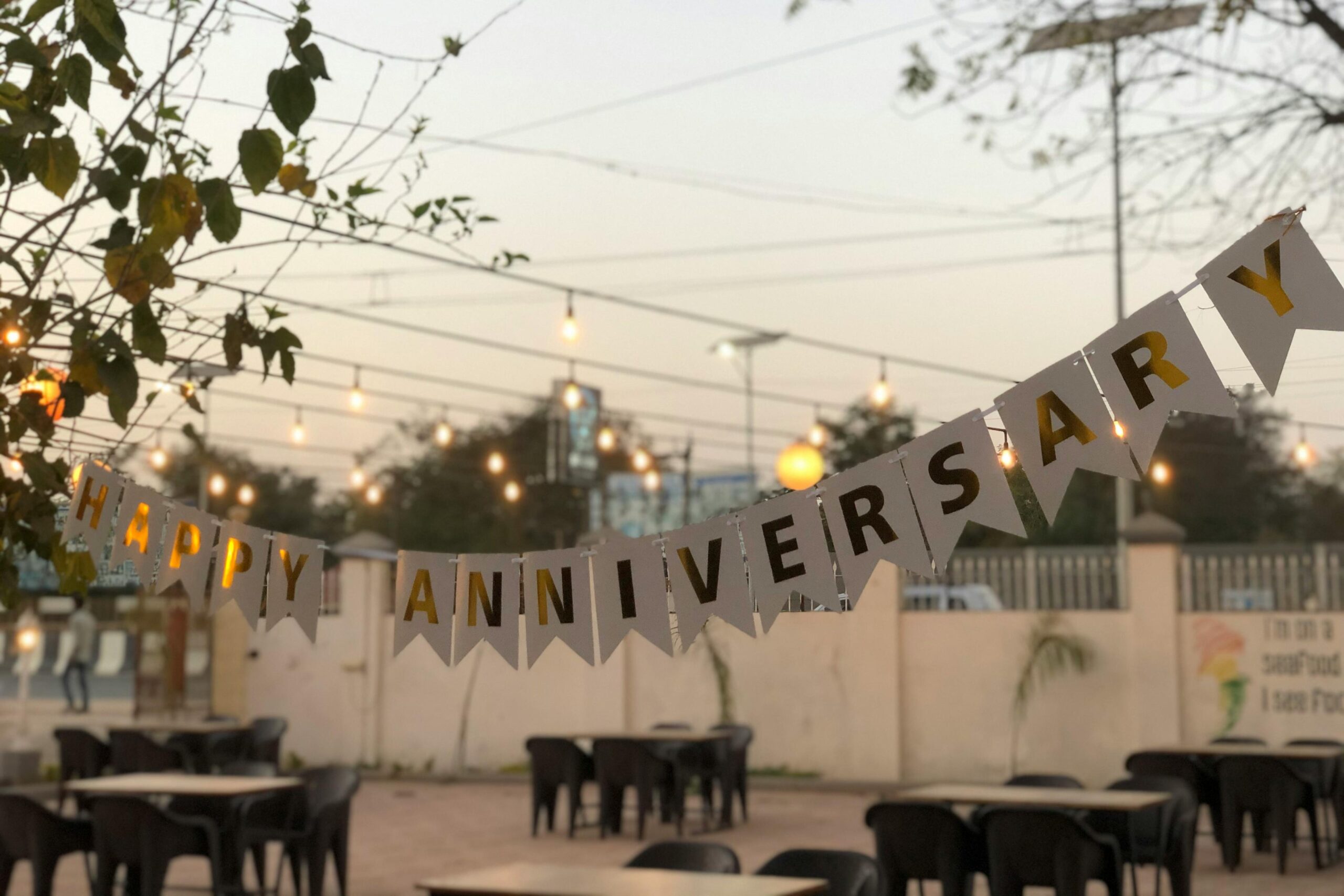A white "HAPPY ANNIVERSARY" banner with gold lettering hangs across an outdoor patio set with several dining tables and chairs. The background features glowing string lights and a soft evening sky.