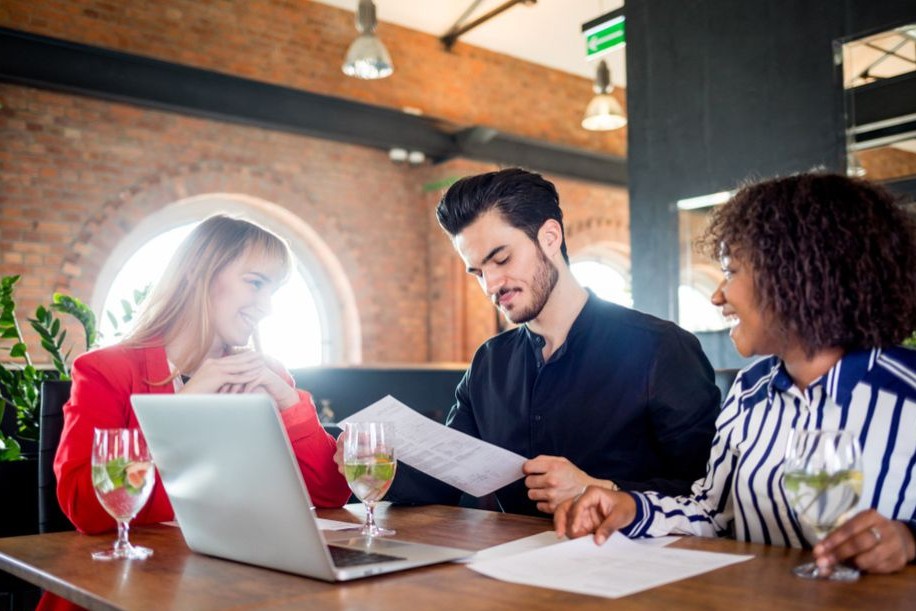 Three people sit around a wooden table in a bright, industrial-style restaurant while reviewing documents and looking at a laptop. They appear to be engaged in a professional meeting or business discussion.