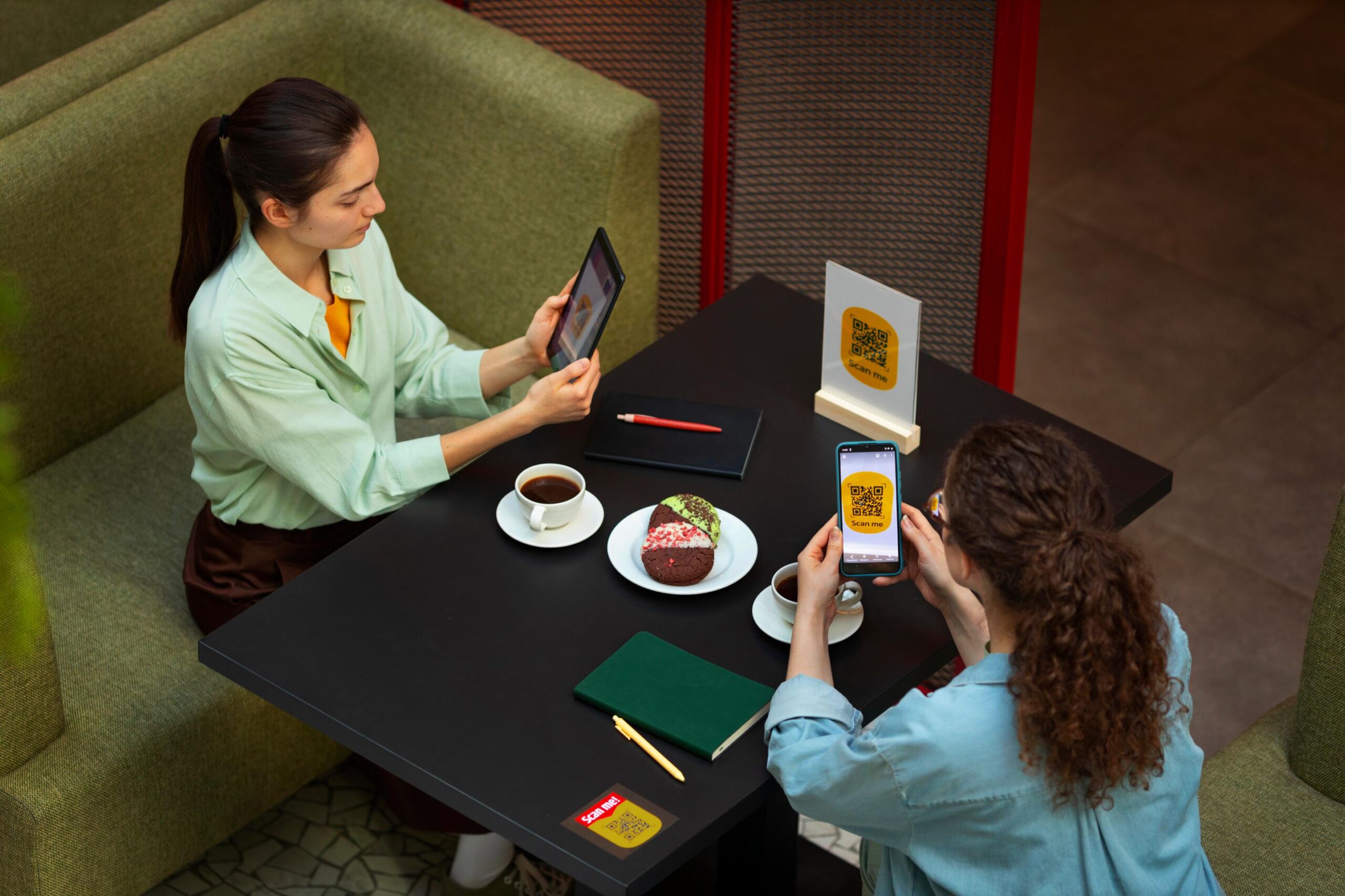 Two women sit at a cafe table, using a tablet and a smartphone to scan QR codes for a digital menu. Between them, the table is set with cups of coffee, a plate of colorful cookies, and two closed notebooks with pens.