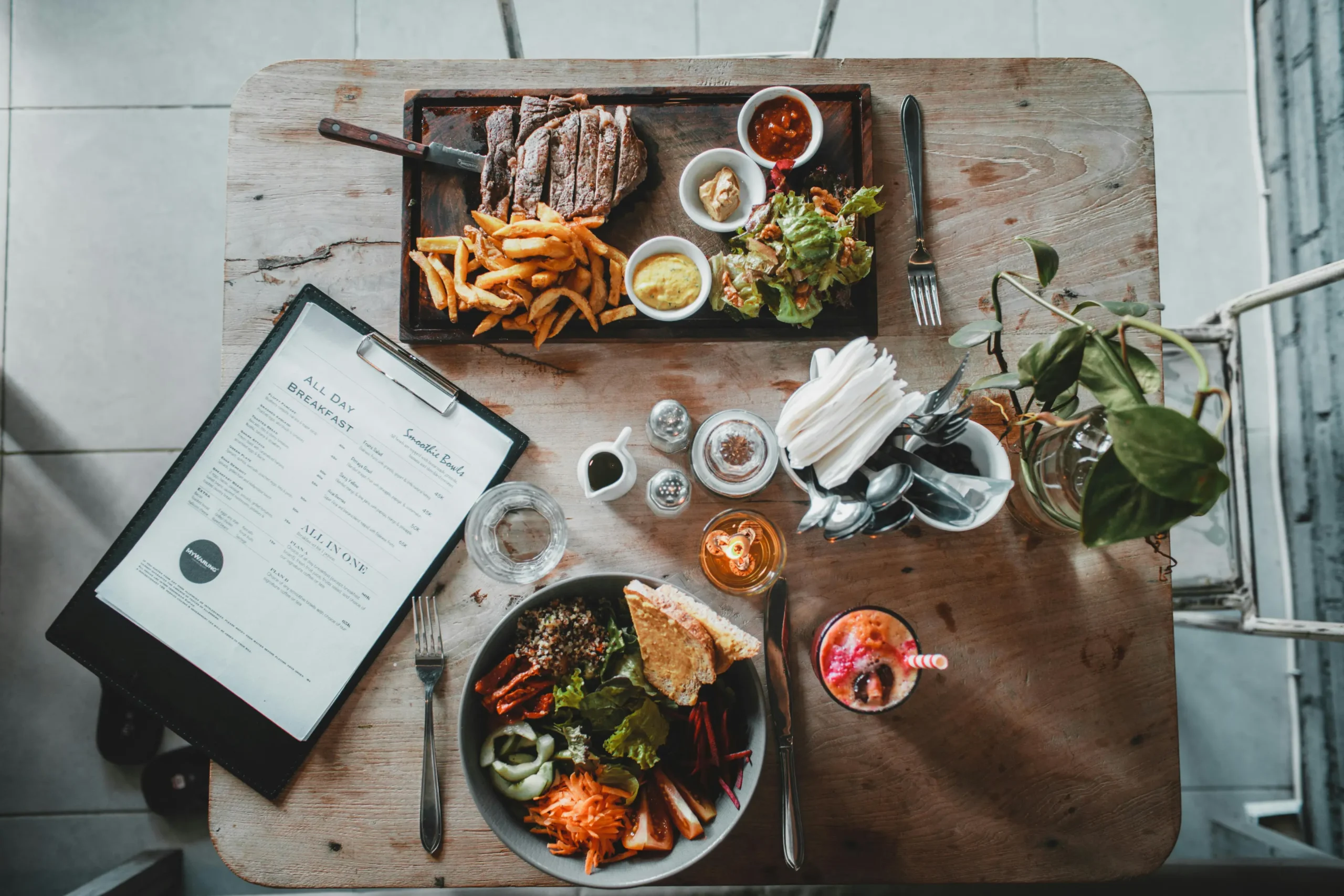 A rustic wooden table is spread with a variety of breakfast and lunch dishes, including steak with fries and a fresh vegetable grain bowl.