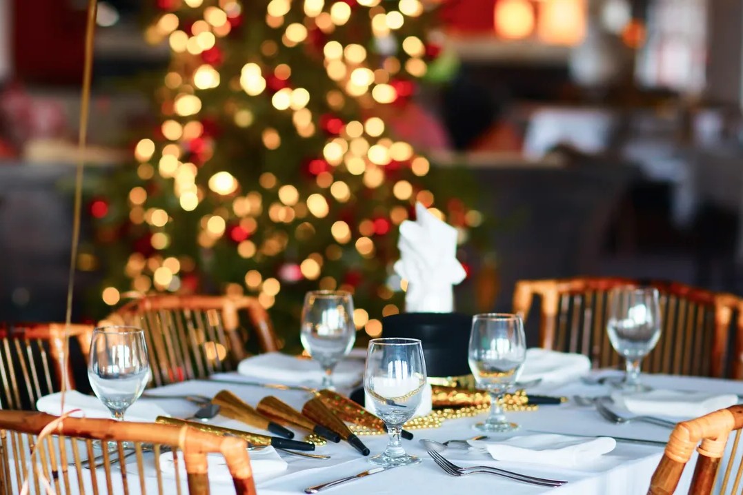 A festive dining table is elegantly set with white linens, wine glasses, and gold party horns in preparation for a celebration. In the background, a beautifully blurred Christmas tree glows with warm, shimmering bokeh lights.
