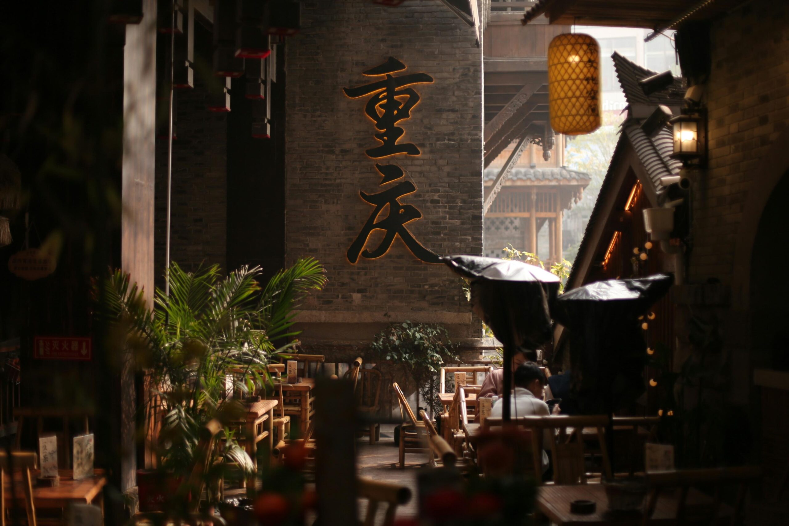 This atmospheric photo shows a traditional Chinese courtyard café featuring bamboo furniture and a glowing yellow lantern. Large, backlit calligraphy characters meaning "Chongqing" are prominently displayed on a dark brick wall in the background.
