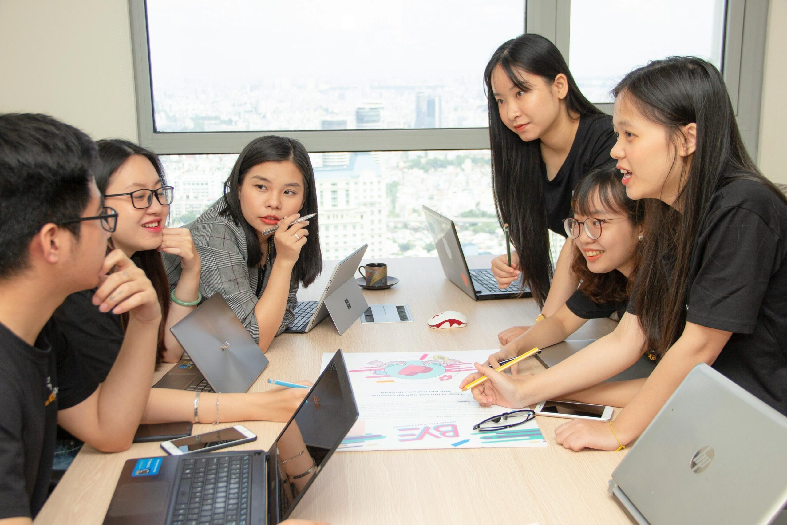 A group of young professionals is gathered around a conference table in a bright office, engaging in a collaborative discussion over a colorful printed document. Several open laptops and smartphones are scattered across the table.