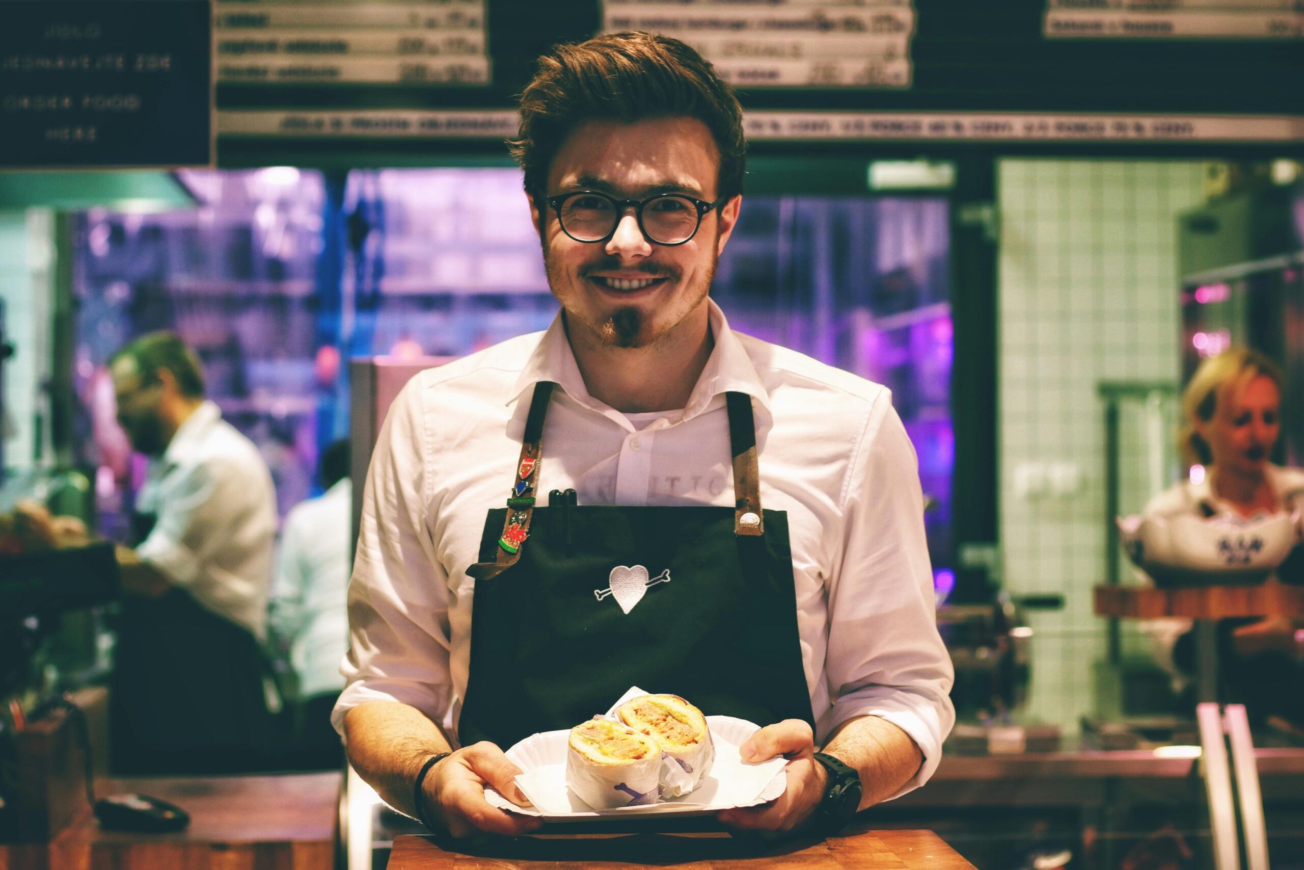 A smiling man wearing glasses and a dark apron stands behind a counter, proudly presenting a halved sandwich on a white paper plate.