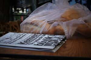 A folded Turkish newspaper and a large plastic bag filled with fresh bread sit atop a wooden table. The scene captures a quiet, everyday moment, likely at an outdoor cafe or a local market.