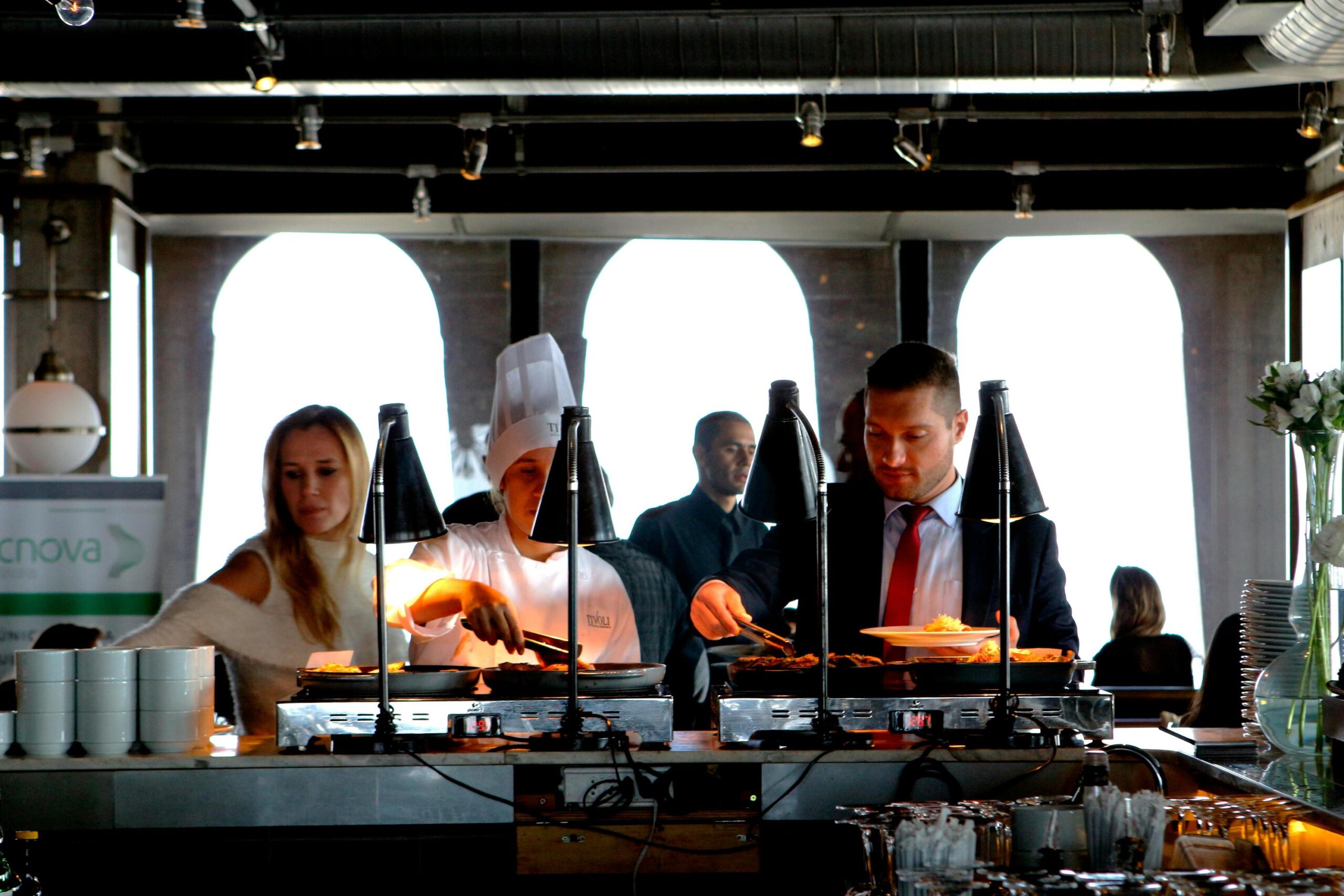 A chef in a tall white hat and a man in a suit serve themselves from a buffet line under warm heat lamps. The background features large arched windows and several other guests in a brightly lit, modern dining space.