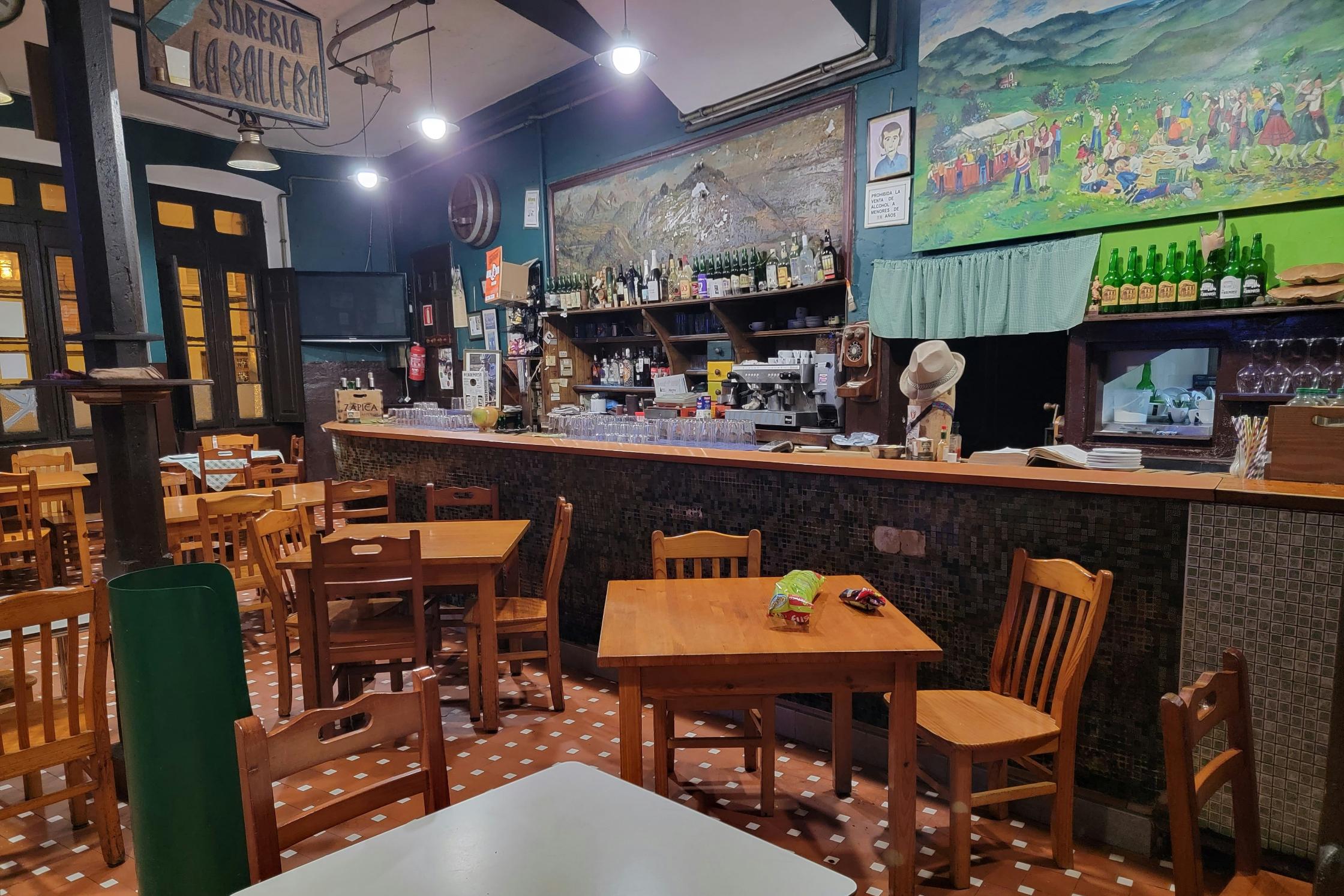 The image showcases the rustic interior of a traditional Spanish cider house, or sidrería, featuring dark teal walls, a tiled bar, and simple wooden tables. Colorful folk art and rows of green bottles decorate the space.
