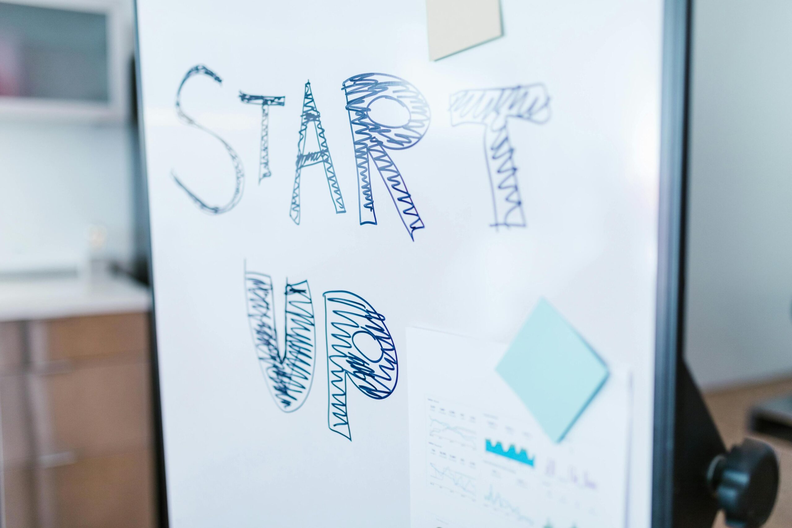 A whiteboard features the words "START UP" hand-drawn in large, blue-inked block letters. Attached to the board are colorful sticky notes and a printed document displaying various data charts and graphs.