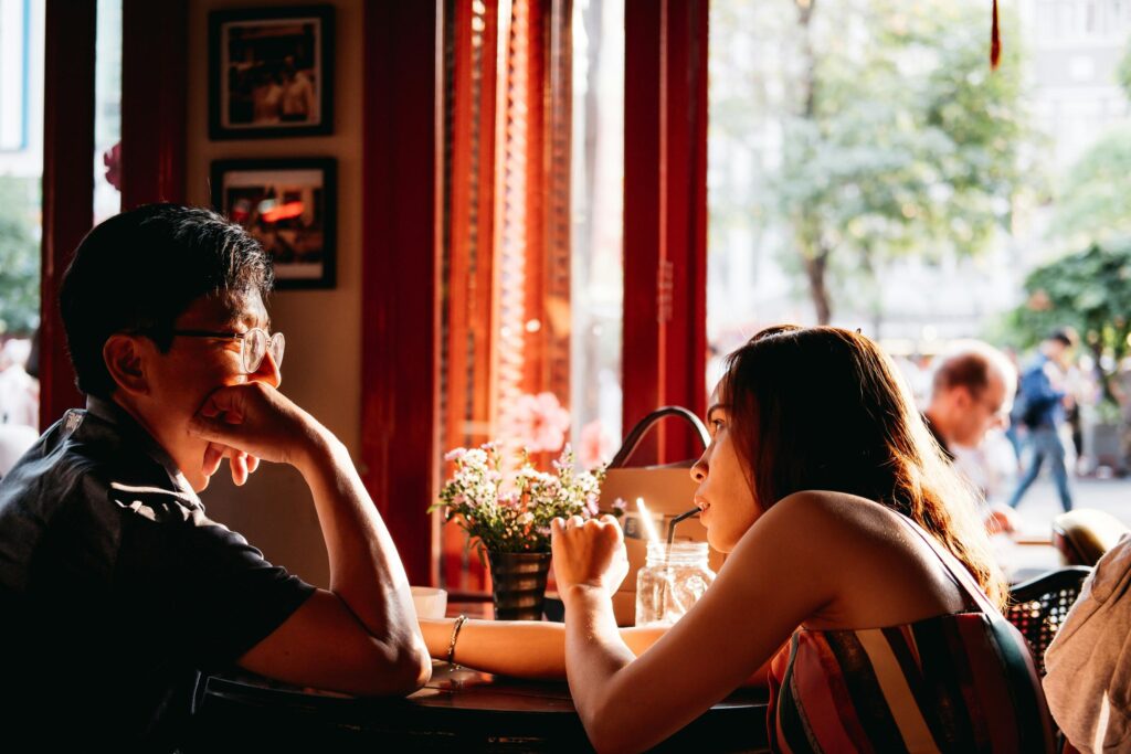 wo people are seated at a circular table in a sunlit cafe, engaged in an intimate conversation near a large window. A small vase of flowers and a mason jar drink sit between them, highlighted by the warm a glow.