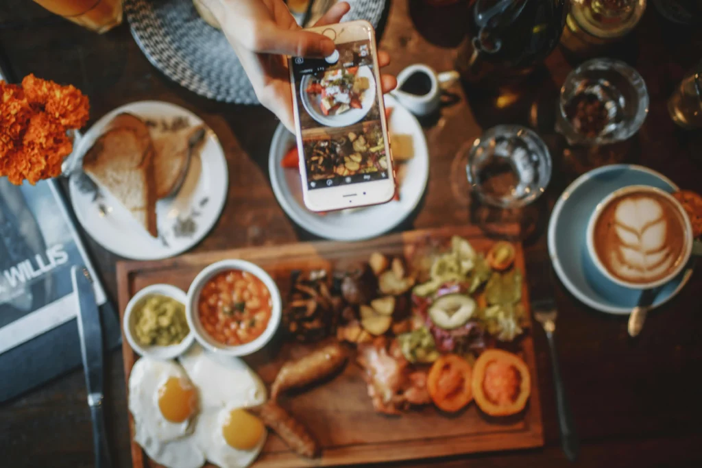 A person holds a smartphone over a rustic wooden table to capture a flat-lay photo of a hearty breakfast spread. The meal includes fried eggs, sausages, beans, and toast, accompanied by a latte with latte art.