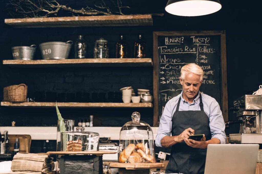 An older man in a black apron stands behind a cafe counter, focused on using a small handheld calculator or device. Behind him, rustic wooden shelves and a chalkboard menu create a cozy, industrial atmosphere filled with pastries and coffee equipment.