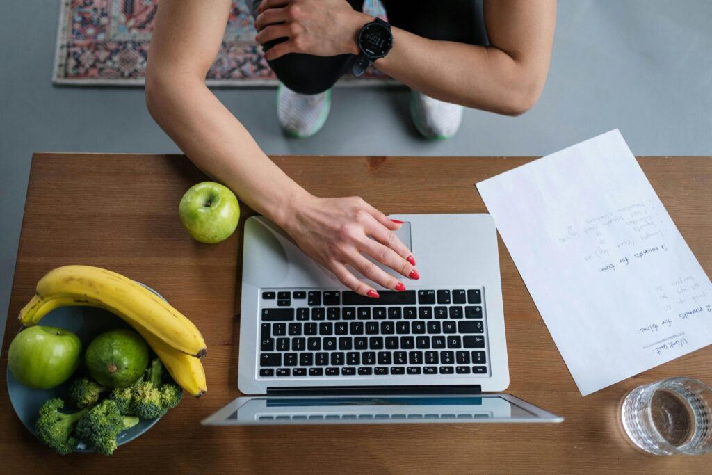 A person in workout gear uses a laptop on a wooden table surrounded by a bowl of fruit, vegetables, and a handwritten fitness plan.