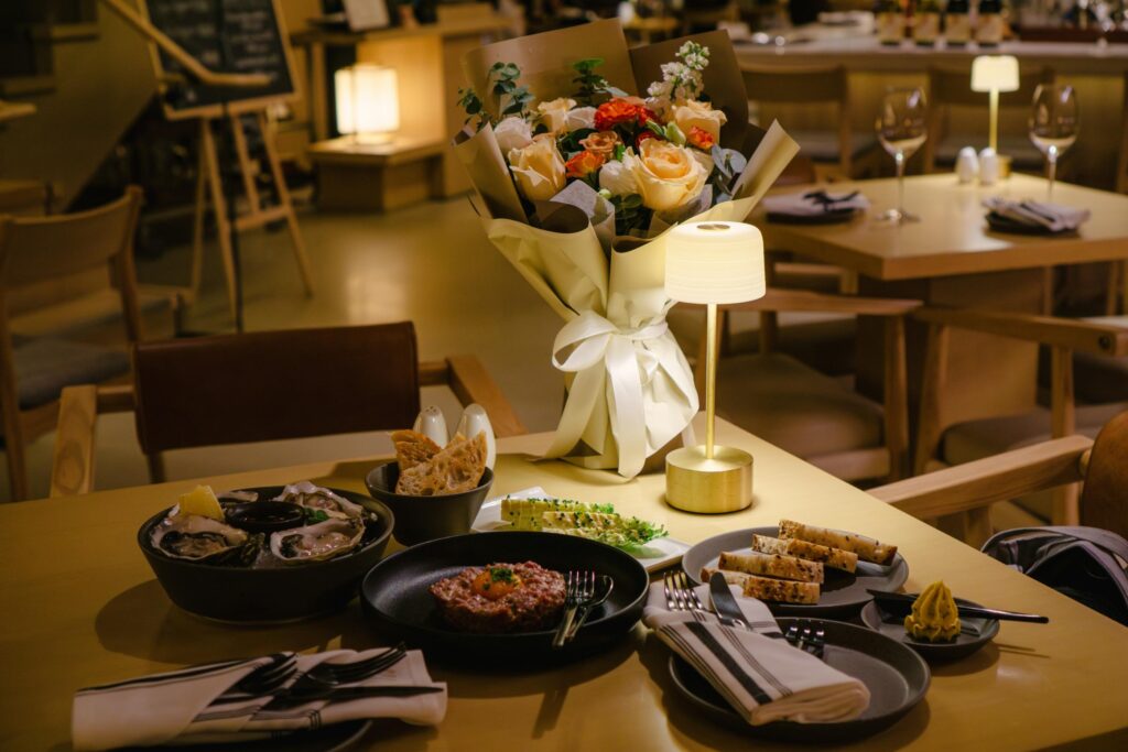 A candlelit dinner table is set with a bouquet of flowers, oysters, steak tartare, and various breads in a warm, dimly lit restaurant. The cozy atmosphere is enhanced by soft lighting from a small desk lamp.