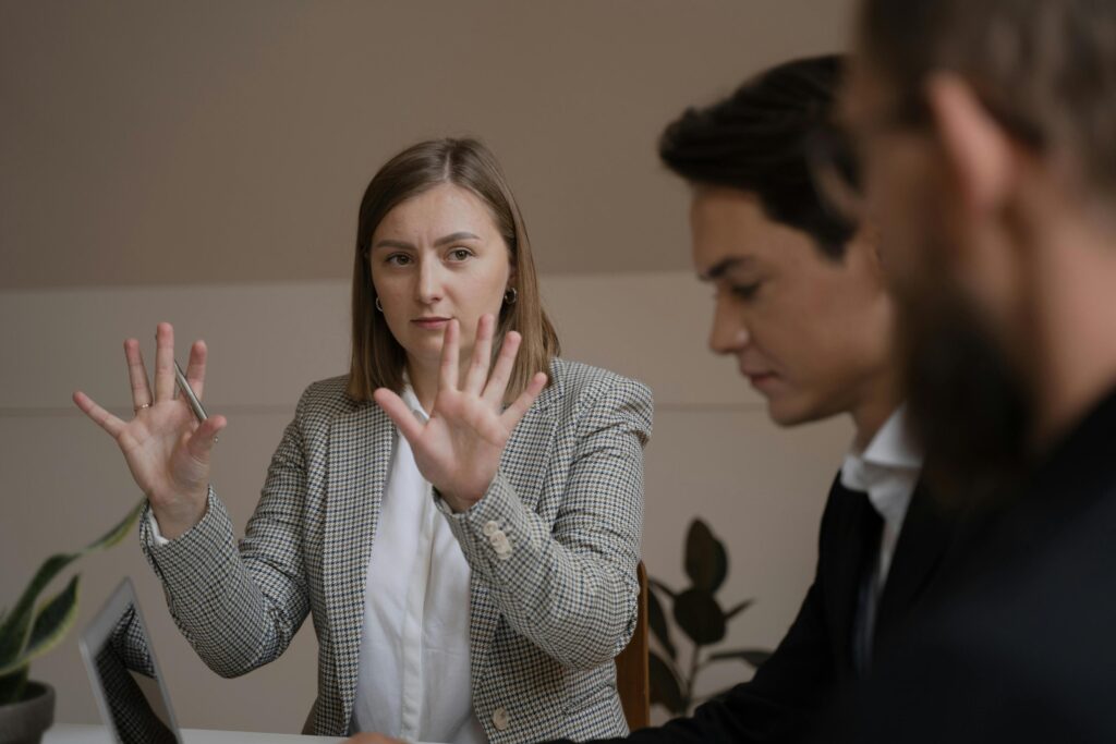 A woman in a patterned blazer holds her hands up in a guiding gesture while addressing two colleagues at a table. The scene captures a focused professional setting.