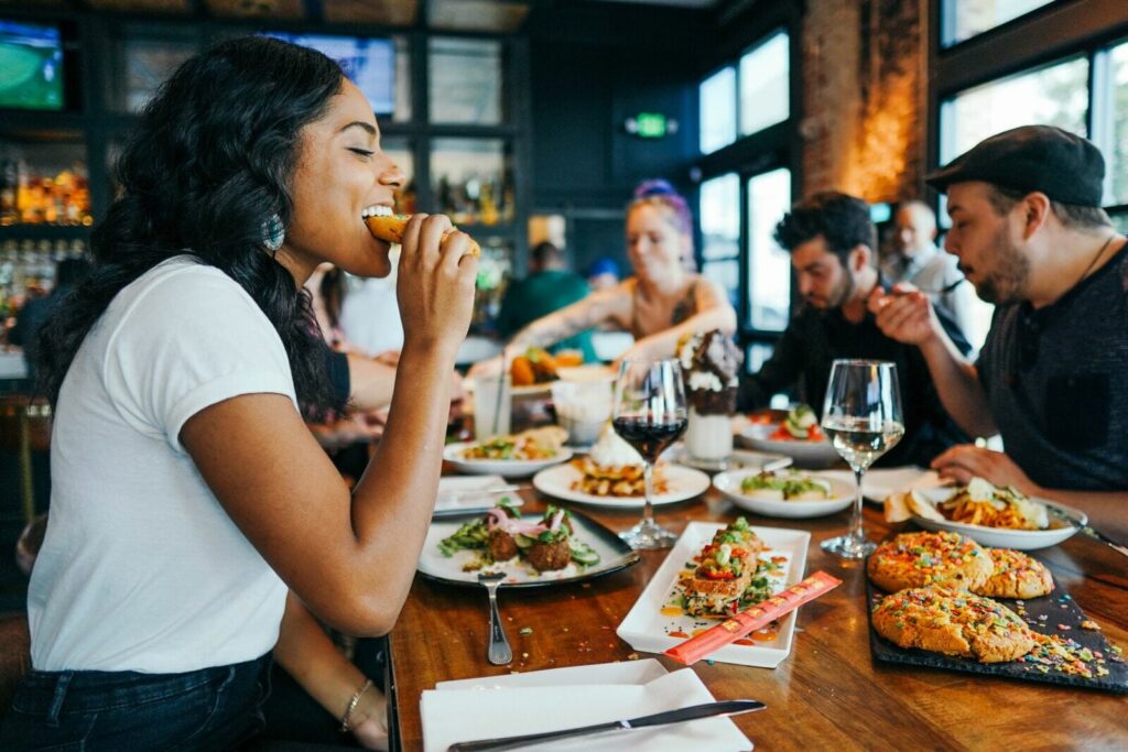 A group of friends enjoys a vibrant meal together at a dimly lit, modern restaurant filled with various small plates and wine. In the foreground, a woman in a white t-shirt smiles as she takes a bite of her food.