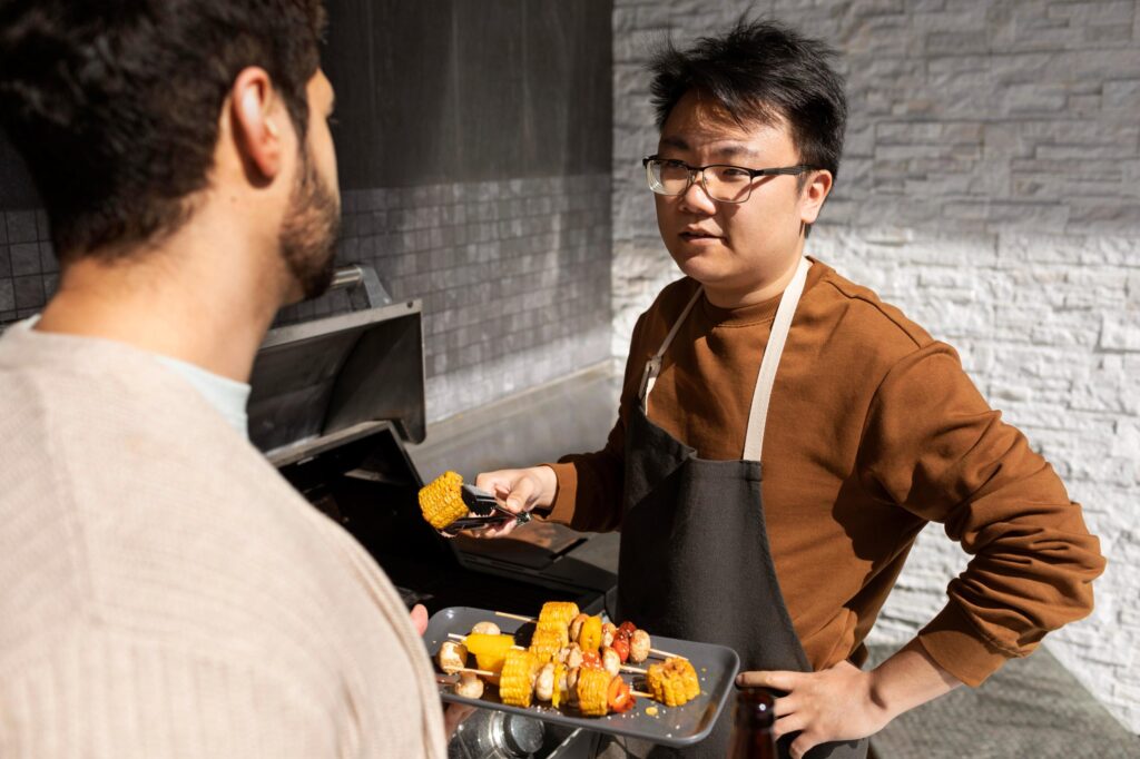 A man in an apron stands by a grill, holding tongs with corn and looking toward his friend. The other man holds a platter of colorful vegetable skewers featuring corn, peppers, and mushrooms.