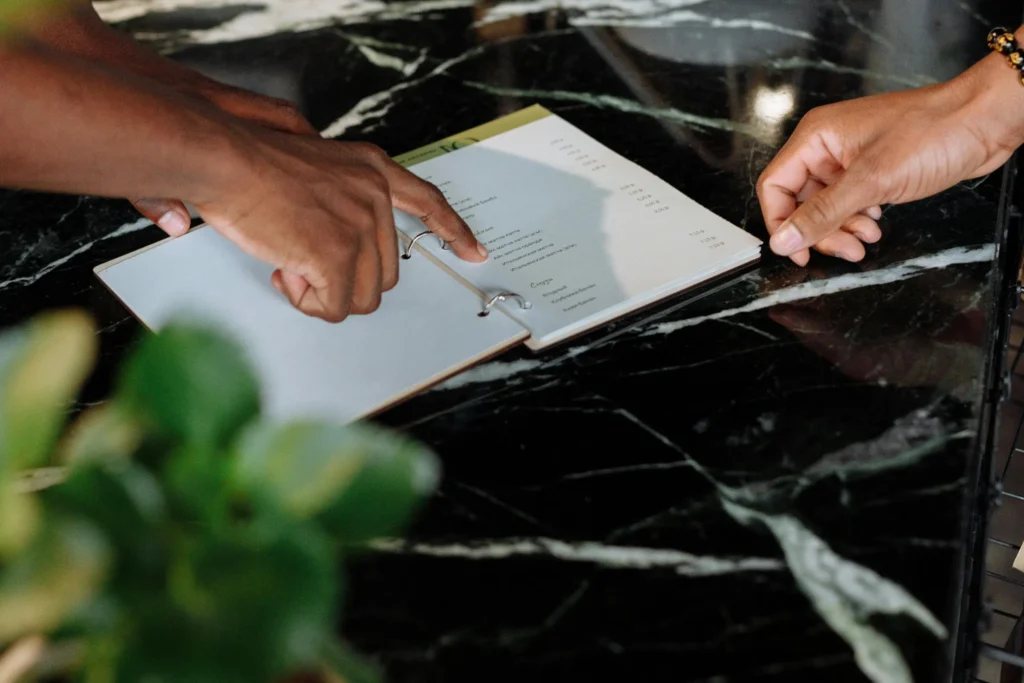Two hands rest on a dark marble countertop while pointing to items in an open, ring-bound menu.