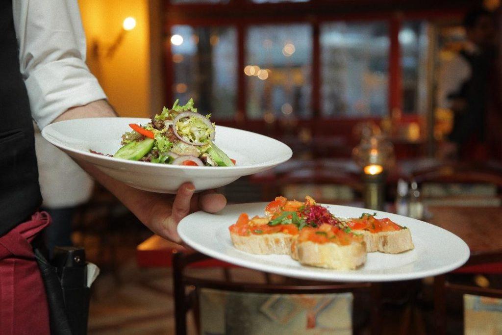 A server holds two white plates of food in a warm, dimly lit restaurant setting. The top plate contains a fresh green salad with cucumbers and red onions
