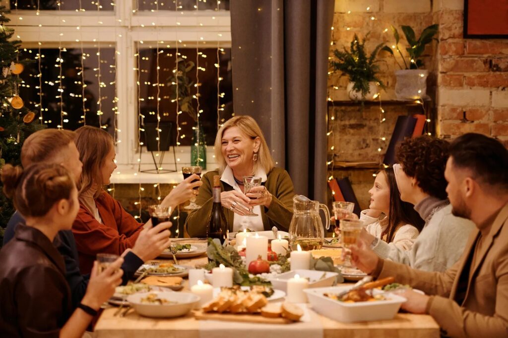 A multi-generational family shares a warm, candlelit meal around a table filled with various dishes and drinks. Glowing string lights and a festive atmosphere create a cozy, celebratory mood for their winter gathering.
