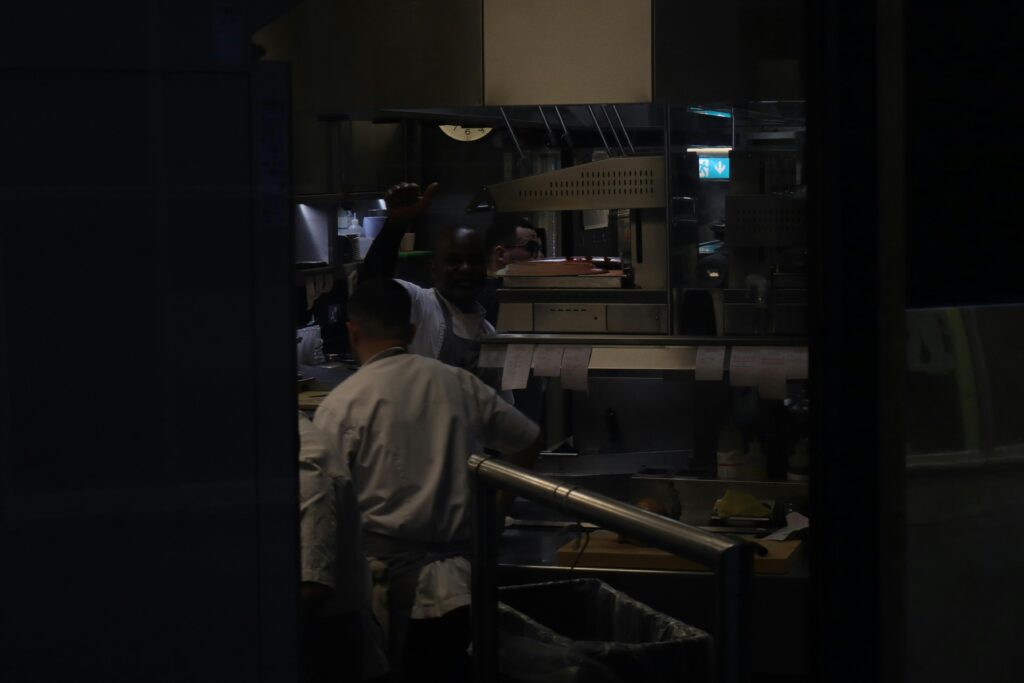 A chef gives an enthusiastic thumbs-up from the center of a busy, industrial kitchen. The candid shot captures a moment of camaraderie amidst the stainless steel equipment and hanging order tickets.