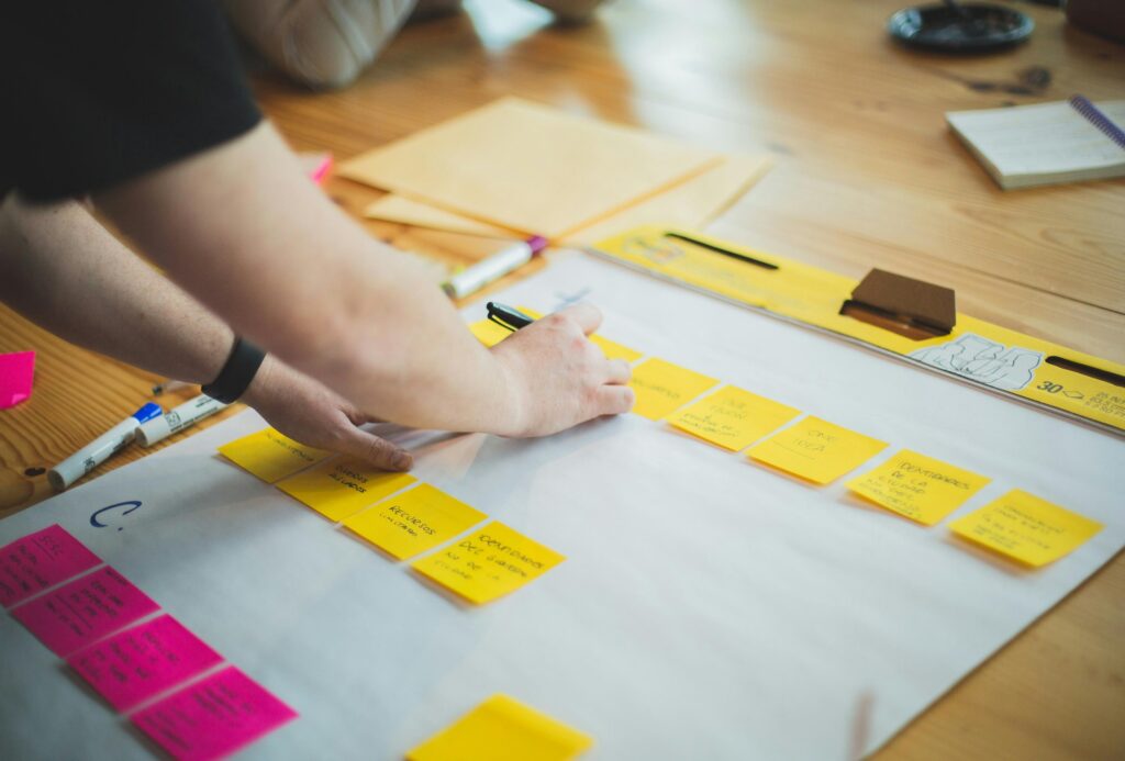 A person’s hands are shown mid-brainstorm, writing on a yellow sticky note placed upon a large white planning sheet. The wooden table is filled with colorful markers and rows of pink and yellow notes, capturing a creative workshop or strategy session in progress.