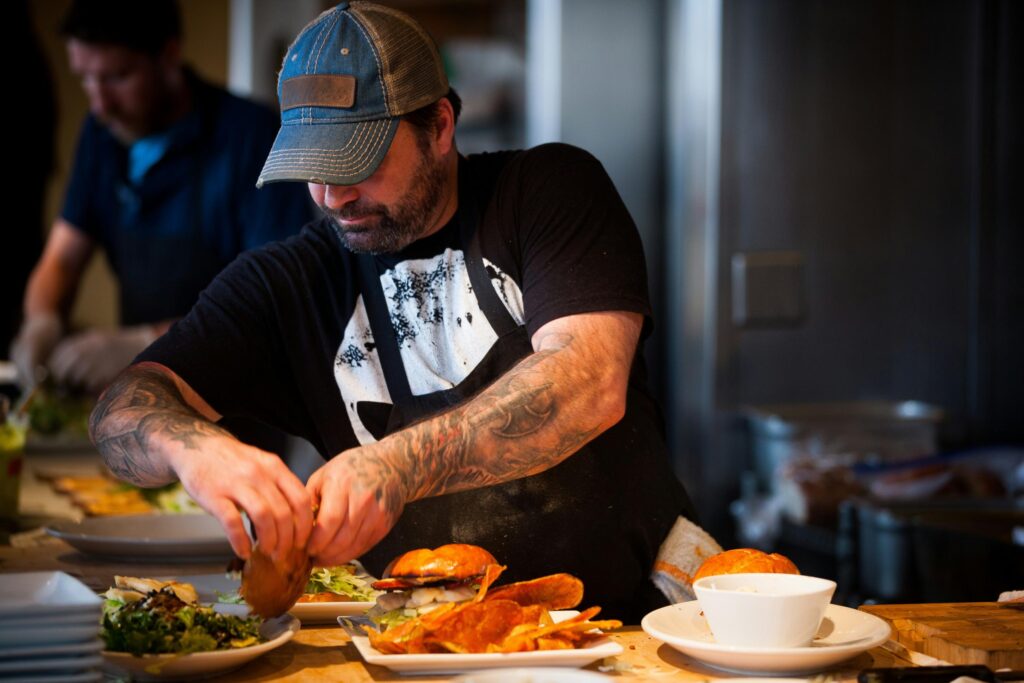 A tattooed man in a baseball cap and apron is focused on assembling gourmet burgers and plating sides in a busy kitchen.