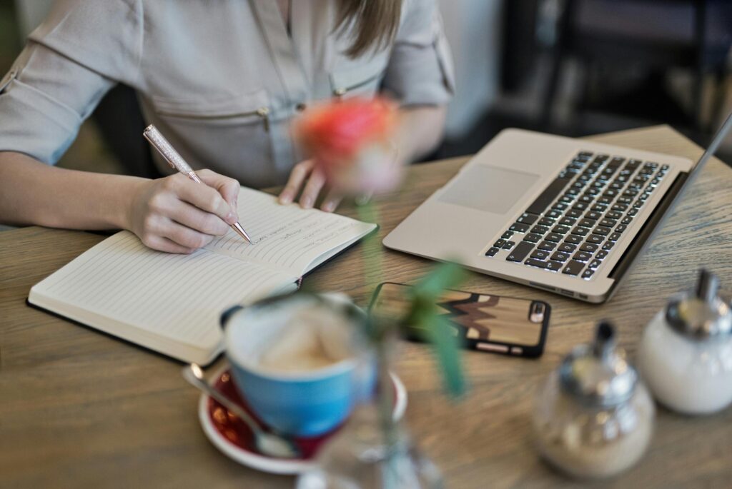A person sits at a wooden table, writing in a notebook next to an open laptop. The cozy workspace is cluttered with a cup of coffee, a smartphone, and a small vase with a flower.