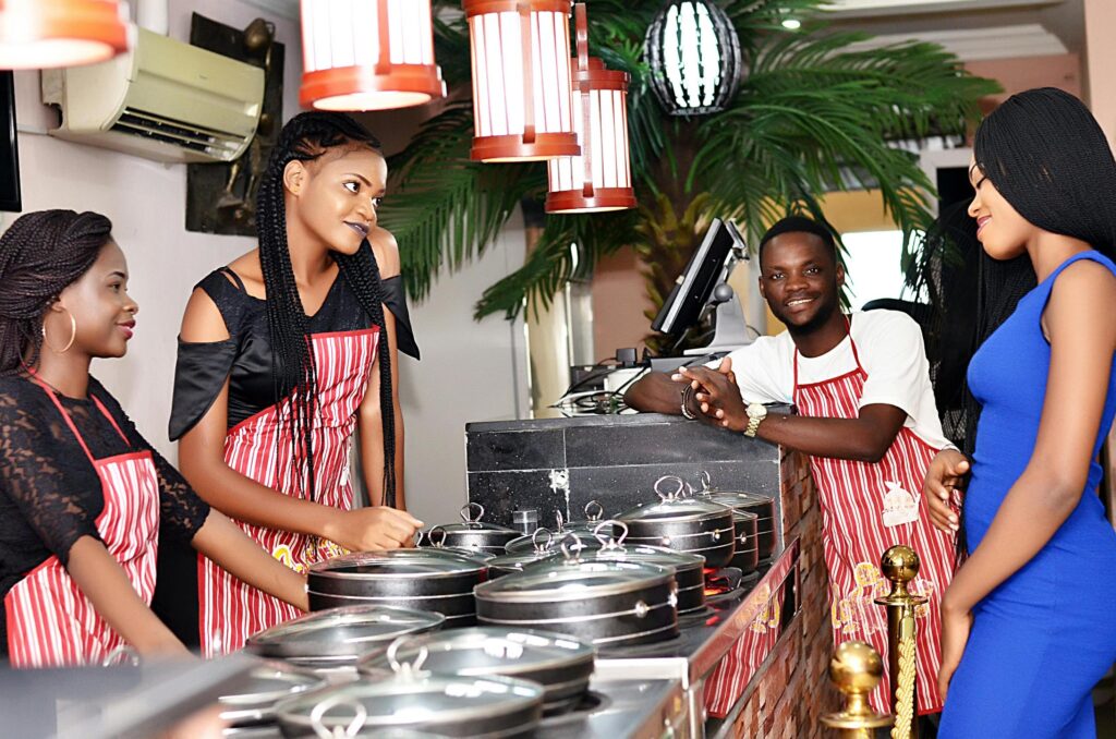 Three staff members in red-and-white striped aprons stand behind a food counter filled with covered pots, interacting with a woman in a blue dress.