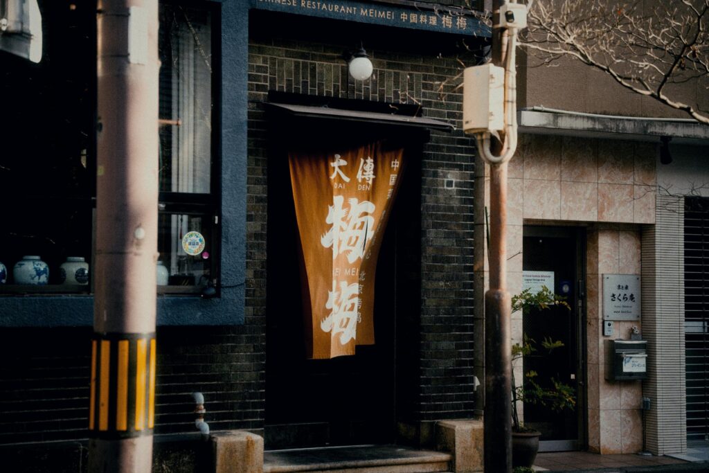 An orange fabric noren hangs over the entrance of a Chinese restaurant titled "Meimei," which is set within a dark brick facade. To the left, a utility pole with yellow and black stripes stands near a window displaying traditional ceramic jars.