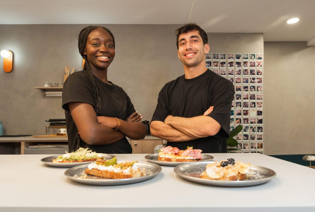 Two smiling individuals stand behind a counter with their arms crossed, showcasing four different types of artisanal toast on metal plates. The background features a minimalist cafe interior with a grid of polaroid photos and warm lighting on the wall.