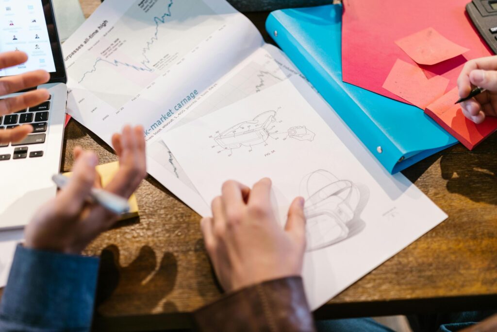 A team is gathered around a wooden table covered in market reports, colorful folders, and product design sketches. They are actively collaborating, with hands gesturing over the documents and a laptop open to a video call.